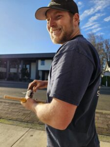A man in a navy T-shirt and cap smiles while holding a wooden stick and a can outdoors on a sunny day, with buildings and trees in the background.