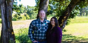 A man and woman stand side by side outdoors in front of trees and grass, smiling at the camera on a sunny day.