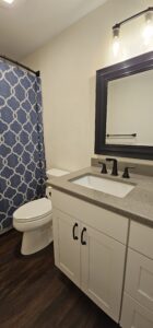 A modern bathroom with a white vanity, gray countertop, black fixtures, a toilet, dark wood floor, and a blue shower curtain with a white geometric pattern.