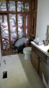 A person is working on plumbing near a toilet in a partially demolished bathroom with exposed wall insulation and construction tools on the counter.