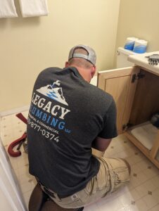 A plumber wearing a Legacy Plumbing t-shirt kneels on a bathroom floor while working under a sink cabinet.