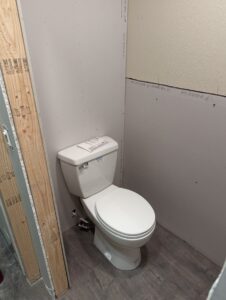 A white toilet installed in a partially finished bathroom with exposed drywall and wood framing, and a laminate wood floor.