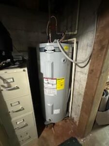 A gray water heater is installed in a basement corner next to a filing cabinet, with pipes connected at the top and warning labels visible on the tank.