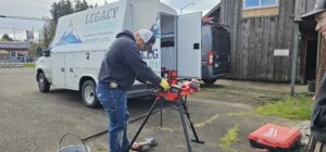 A plumber wearing a cap and gloves works with tools on a portable stand next to a Legacy Plumbing van parked outside a wooden building.