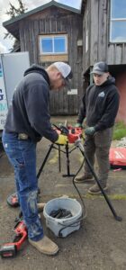 Two people wearing work clothes operate a pipe threading machine on a tripod outdoors, with a wooden building and tools visible in the background.