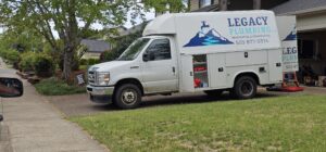 A white Legacy Plumbing service van is parked in a residential driveway next to a house and trees, with equipment visible in the open garage.