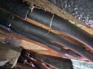 Black foam pipe insulation is installed along wooden floor joists in a crawl space, with some cobwebs visible hanging from the wood.
