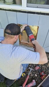 A man uses a reciprocating saw to cut a square opening in the exterior wall of a house, exposing yellow insulation. Tools are scattered on the ground nearby.