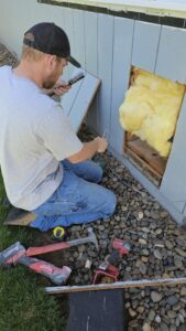 A man repairs the exterior wall of a building, exposing insulation and using tools including a drill, tape measure, and hammer, while kneeling on rocks.