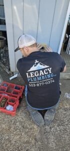 A plumber in a Legacy Plumbing shirt works on an exterior wall beside a red toolbox filled with various tools and hardware.