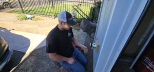 A person wearing a cap and black shirt kneels by a door, working on the doorknob with tools and a toolbox nearby.