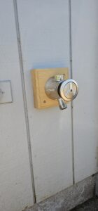 A metal outdoor water spigot with a locking device is mounted on a wooden block attached to a white wall.