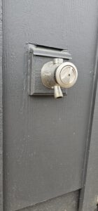 A metallic outdoor faucet with a locking mechanism is mounted on a dark gray wooden wall.