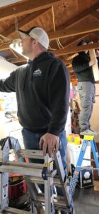 Two men working in a garage; one stands on a ladder reaching toward the ceiling, while the other holds a wooden board and stands on a step stool in the background.