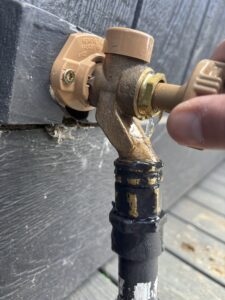 A close-up of a hand turning an outdoor faucet, with water leaking from the connection point above a black-painted pipe.