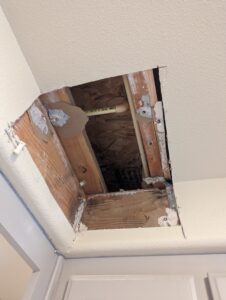 A rectangular hole cut into a ceiling reveals wooden beams, pipes, and insulation materials inside the ceiling cavity.