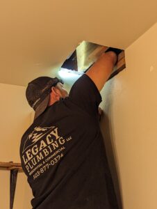 A plumber in a Legacy Plumbing shirt works with one arm extended into a hole in the ceiling, using a flashlight for visibility.