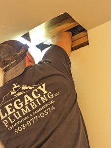 A plumber from Legacy Plumbing repairs pipes through a hole in a ceiling, wearing a company t-shirt with contact information printed on the back.