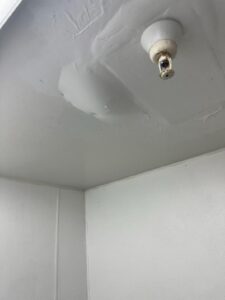 A ceiling with visible water damage and patching near an empty light socket in a corner of a room with white walls.
