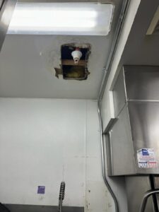 A ceiling with a hole exposing insulation and a light bulb socket, located above a commercial kitchen sink area.