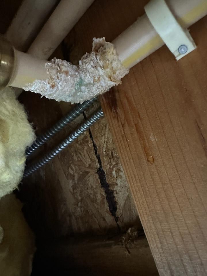 Close-up of a corroded and leaking plastic pipe attached to a wooden surface, with mineral buildup and a small drop of water visible—suggesting repiping may be needed to prevent further damage.