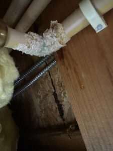A close-up of a pipe with significant corrosion and mineral buildup, next to wood framing and a small water droplet.