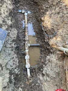 A damaged water pipe is exposed in a muddy trench, with standing water and soil around it. Tools and a pipe fitting are visible nearby.
