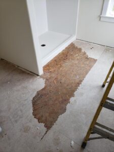 A section of bathroom flooring is partially removed near a shower, exposing the subfloor. A yellow ladder is positioned nearby.
