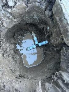A blue PVC pipe fitting in a muddy hole surrounded by wet soil, partially submerged in water.