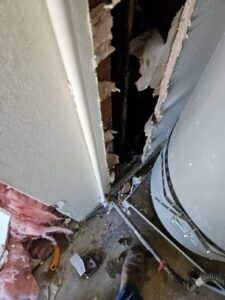 A section of drywall near a water heater is removed, exposing pipes and insulation. Pink insulation and tools are scattered on the floor.