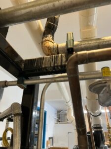A close-up view of intersecting metal pipes, some with rust and corrosion, and others insulated, in an indoor industrial or mechanical room setting.