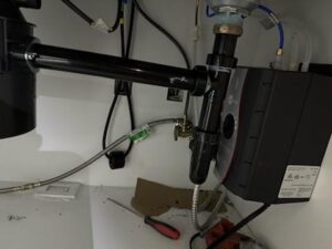 Plumbing and electrical connections under a sink, showing pipes, cables, a black box unit, a screwdriver, and miscellaneous items on the cabinet floor.