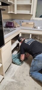 A man kneels on the floor and works under a kitchen cabinet in a partially renovated kitchen with open shelves and no doors.