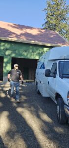 A person wearing work clothes and gloves stands beside a white utility van parked in front of a partially constructed building.
