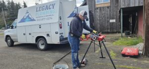 A plumber wearing gloves and a hat uses a pipe threading machine outdoors next to a Legacy Plumbing work van and a wooden building.