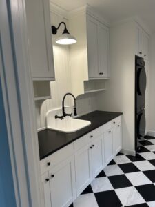 A laundry room with white cabinets, a black countertop, a white sink with a black faucet, stacked washer and dryer, and a black-and-white checkered tile floor.