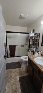 A bathroom with a shower featuring frosted sliding doors, a toilet, a sink with a granite countertop, shelves with toiletries, and gray floor mats.