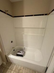 A white bathtub with built-in shelves is installed in a bathroom with beige walls and tiled floor; a box sits inside the tub.