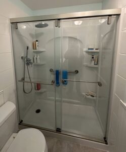 Glass-enclosed shower with sliding doors, grab bars, shelves with toiletries, a rainfall showerhead, and a handheld showerhead; two blue towels hang inside.