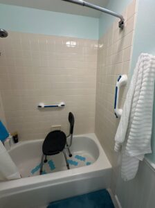A shower chair is placed inside a bathtub with non-slip decals. Grab bars are mounted on the tiled wall, and a white towel hangs on a hook.