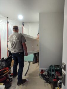 Two workers carry a bathtub into a partially renovated bathroom with tools and construction materials scattered on the floor.