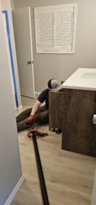 A person wearing gloves and a cap sits on the floor installing or repairing plumbing under a bathroom vanity with tools nearby.