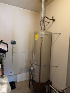 A tall water heater is installed in a corner of a room, with pipes and a vent attached; a digital device is mounted on the nearby wall.