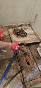 A person wearing red gloves uses a crowbar and hammer to remove damaged wooden floorboards with visible rot and holes.