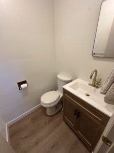 Small bathroom with a toilet, a sink with a cabinet, a wall-mounted mirror, towel, toilet paper holder, and wood-style flooring.
