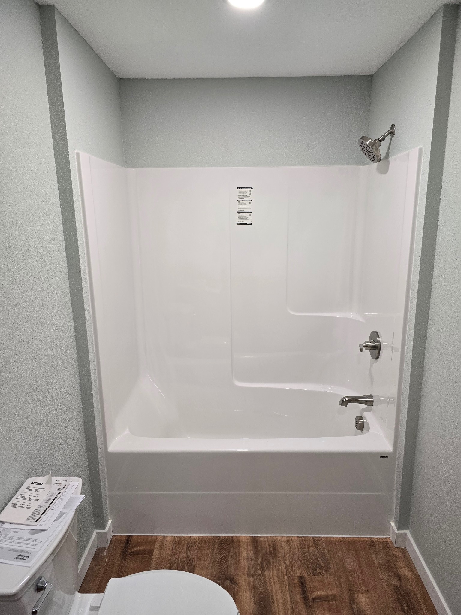 White bathtub and shower combo with a showerhead, faucet, built-in shelves, and water heaters nearby, next to a toilet on a wood-look floor in a small bathroom.