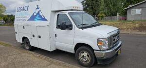 A white Legacy Plumbing service van with a blue logo and contact information is parked on a residential street.