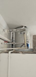 A water heater installed near the ceiling with various pipes and flexible conduits connected, and an expansion tank mounted on top.