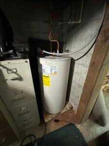 A white water heater is installed in a corner next to a filing cabinet, with pipes connected at the top and a warning label on the front.