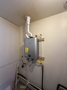 A tankless water heater is mounted on a wall with visible pipes, wiring, and a vent extending to the ceiling in a utility room.